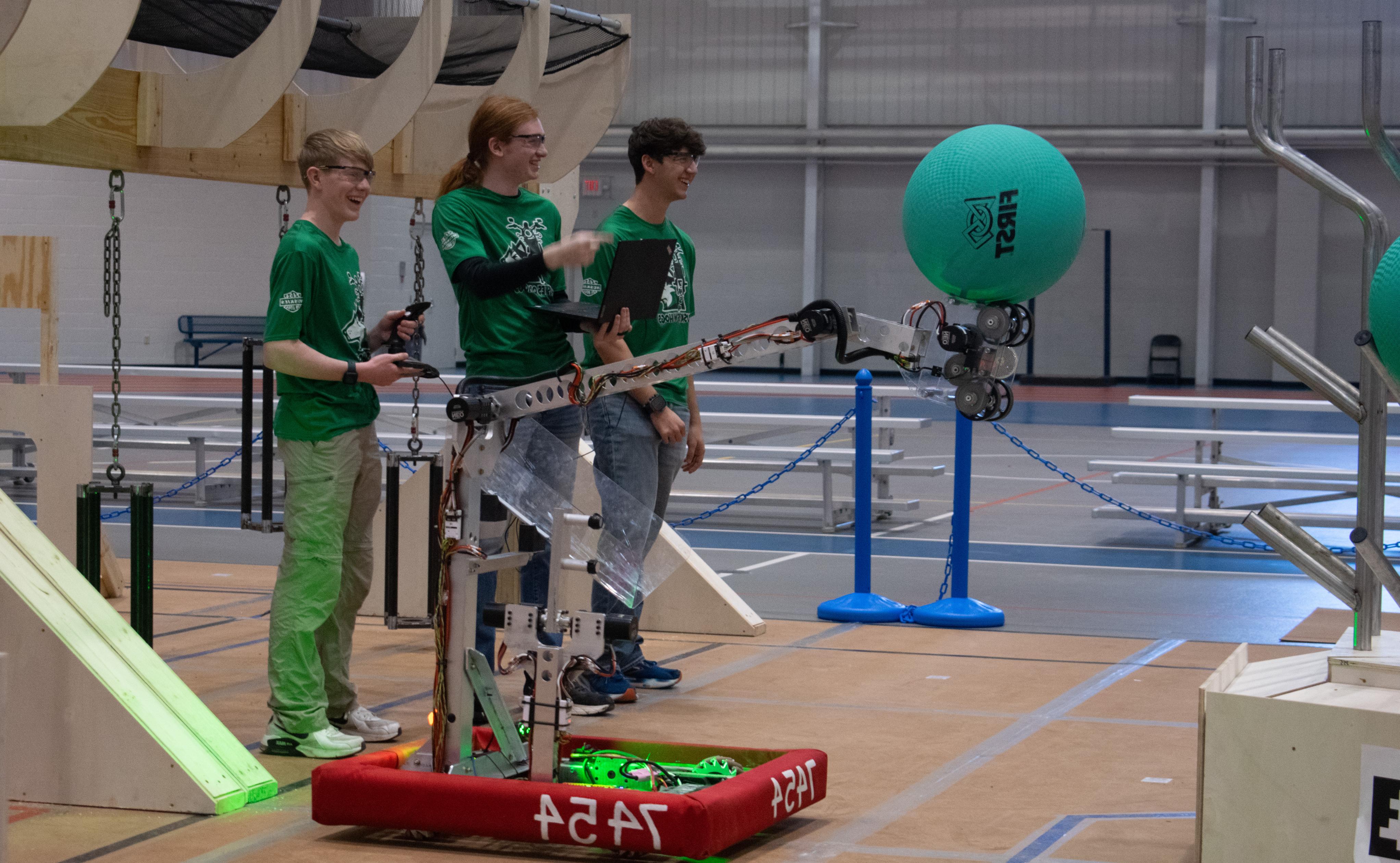 Three male high school students smiling while standing near a robot with an arm extending holding a large ball